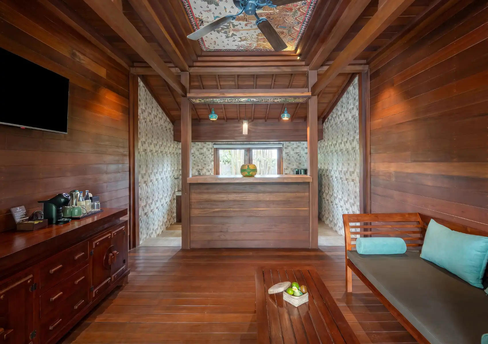 Warm teak wood interior of a Balinese resort villa room featuring a traditional hand-painted ceiling, ceiling fan, Nespresso machine, flat-screen TV, wooden daybed with teal cushions, and a mosaic-tiled en-suite bathroom beyond