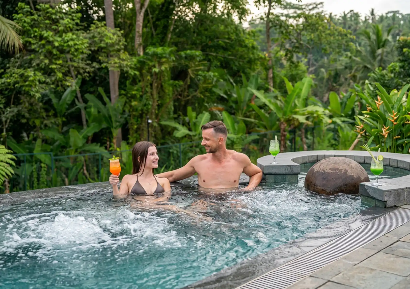 Two people relaxing in a circular outdoor jacuzzi surrounded by tropical greenery, holding colorful drinks with more placed on the stone edge.