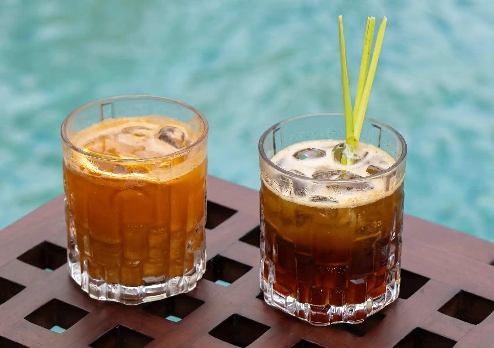 Two iced cocktails served in ribbed rocks glasses on a carved wooden tray beside a turquoise pool in Bali — an amber turmeric-based drink and a dark coffee cocktail garnished with fresh lemongrass stalks