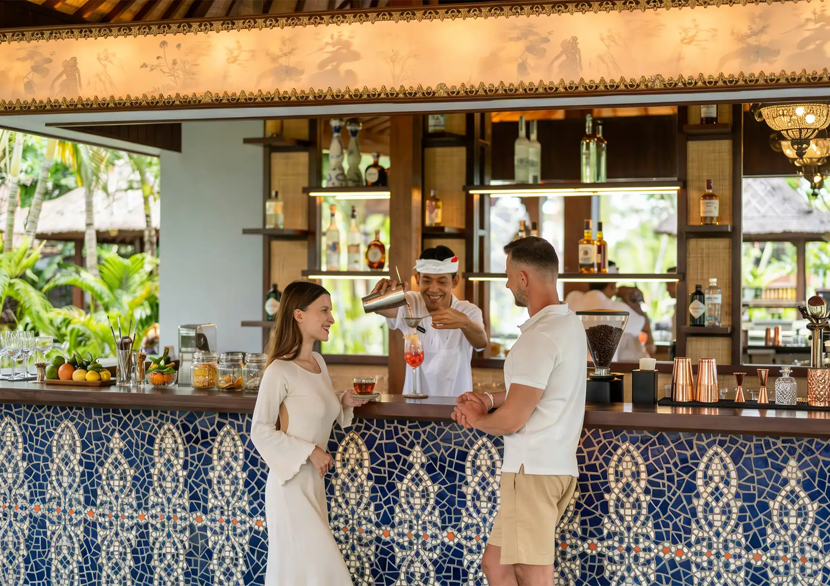 Tropical outdoor bar with blue mosaic counter, bartender pouring a colorful cocktail, and two guests chatting amid lush greenery.