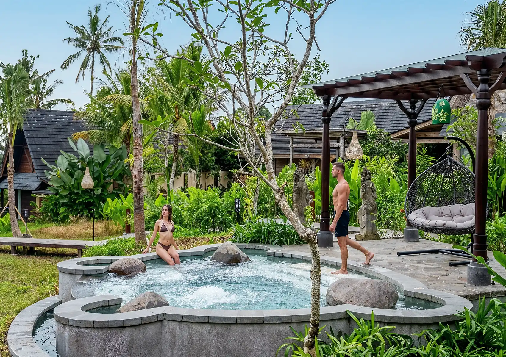 Tropical outdoor spa with circular stone hot tub, lush greenery, and wooden pergola with swing chair near traditional-style buildings.