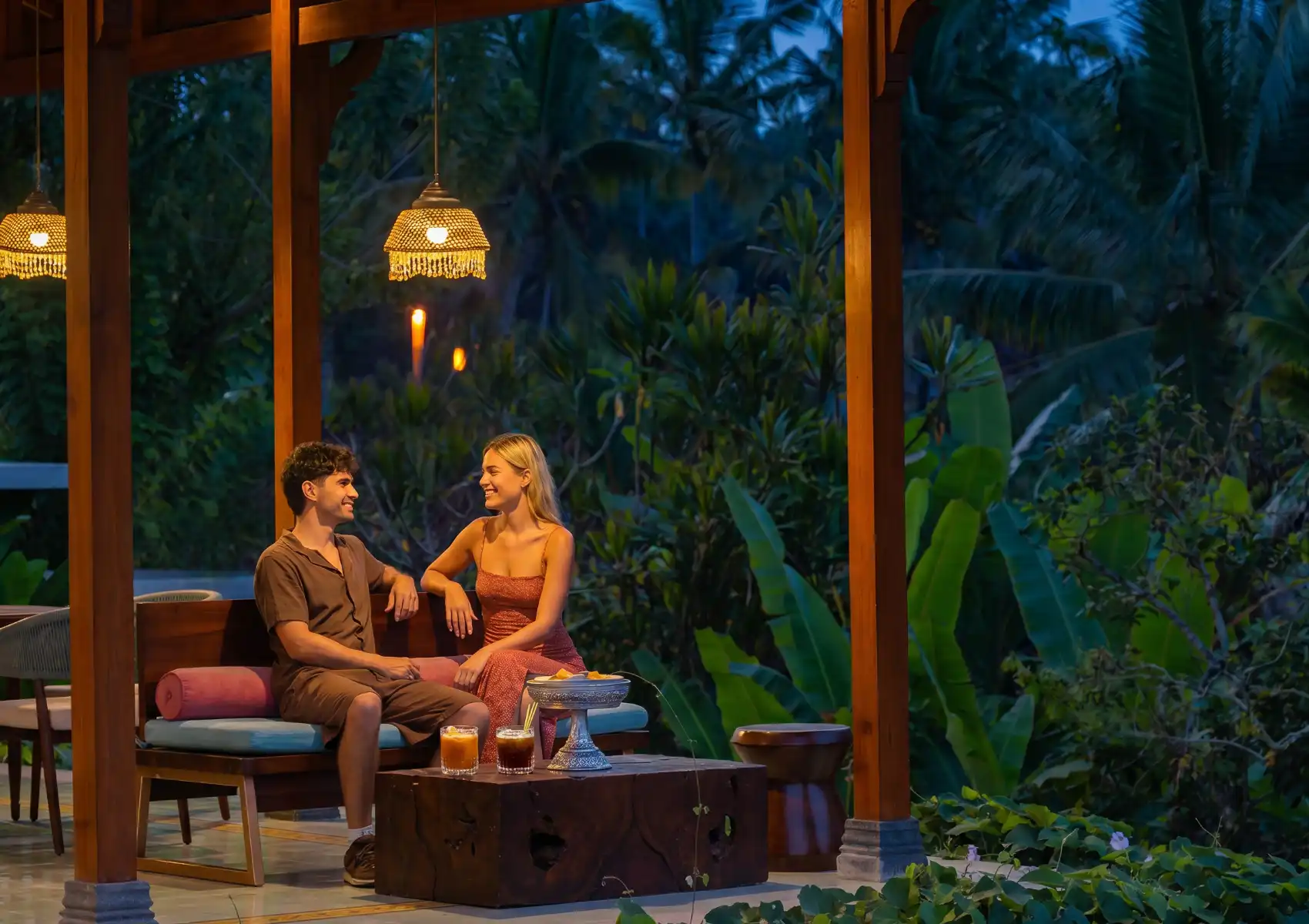 A couple relaxing together on a cushioned outdoor sofa at a candlelit open-air restaurant in Bali at dusk, with iced drinks on a wooden coffee table, beaded pendant lamps, and lush tropical jungle greenery surrounding the pavilion