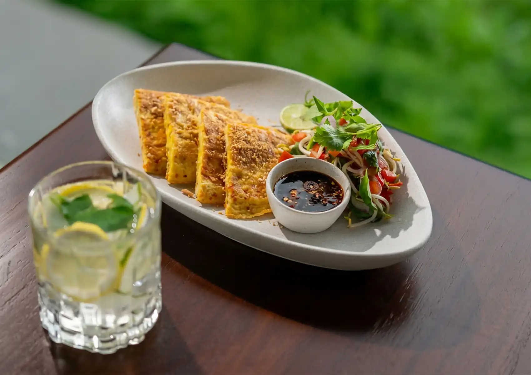 White oval dish with golden-brown savory slices, fresh salad, dipping sauce, and lime wedge, served with iced lemon-mint drink on wooden table.