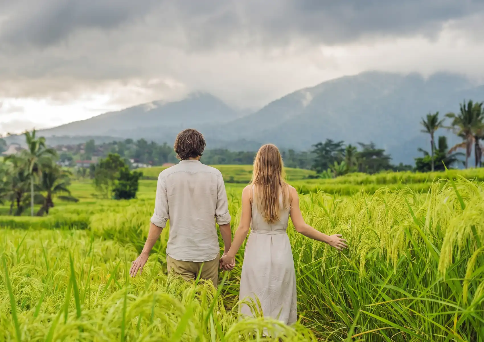A couple holding hands while walking through lush green rice paddies in Ubud, Bali, facing away from the camera toward misty volcanic mountains and dramatic storm clouds on the horizon