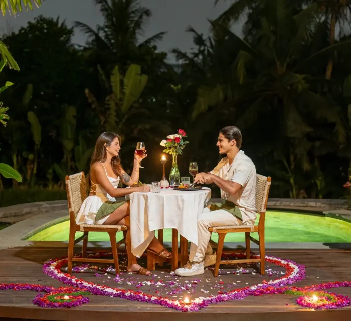 A couple enjoying a romantic Valentine's dinner at Emana Abian Taksu, Ubud, seated at a candlelit white linen table on a teak deck surrounded by heart-shaped flower petal arrangements, tea lights, and a glowing private pool against a tropical night backdrop